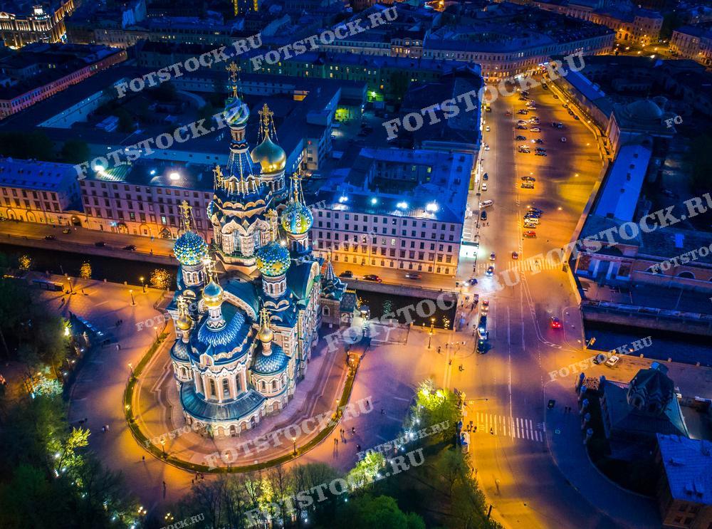 Ночной вид на собор Спаса на Крови в Санкт-Петербурге, снятый с дрона. Фотография подчеркивает яркое освещение и архитектурную красоту собора на фоне городских огней. Идеально подходит для использования в путеводителях, исторических и культурных публикациях.